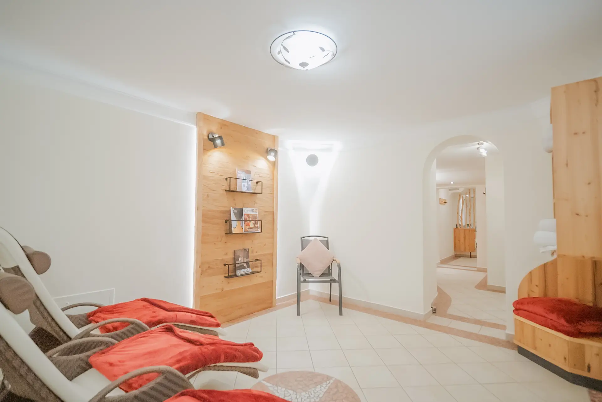 zeiserhof-wellness-11 Heller, moderner Spa-Loungebereich mit Liegestühlen, roten Decken, einem kleinen Holzregal mit Broschüren, weißen Wänden und einer gewölbten Tür, die in einen anderen Raum führt.