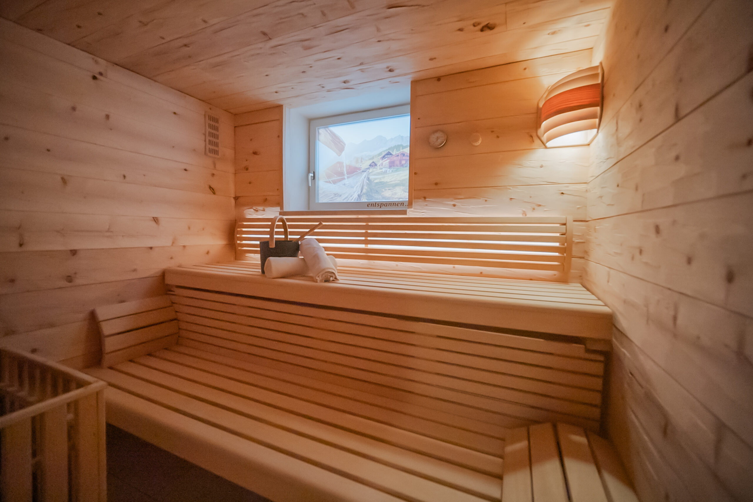 zeiserhof-wellness-3 Der gemütliche Innenraum der Zirbensauna im Zeiserhof verfügt über eine Holzbank, ein Fenster und eine Wandleuchte. Auf der Bank liegen eine Sanduhr und ein Handtuch und schaffen eine einladende Atmosphäre zum Wohlfühlen.