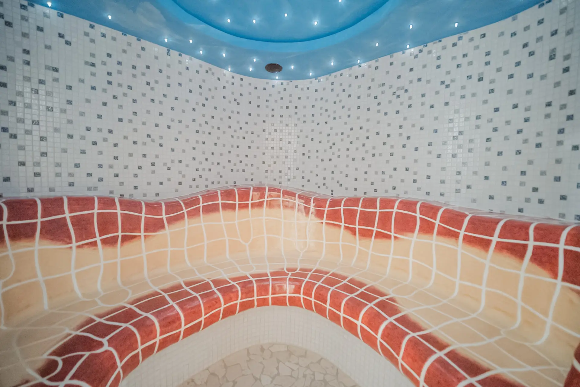 zeiserhof-wellness-5 Ein gefliestes Dampfbad mit einer geschwungenen Bank in Rot und Beige, weißen und grauen Mosaikwänden und einer blauen Decke mit kleinen Lichtern.