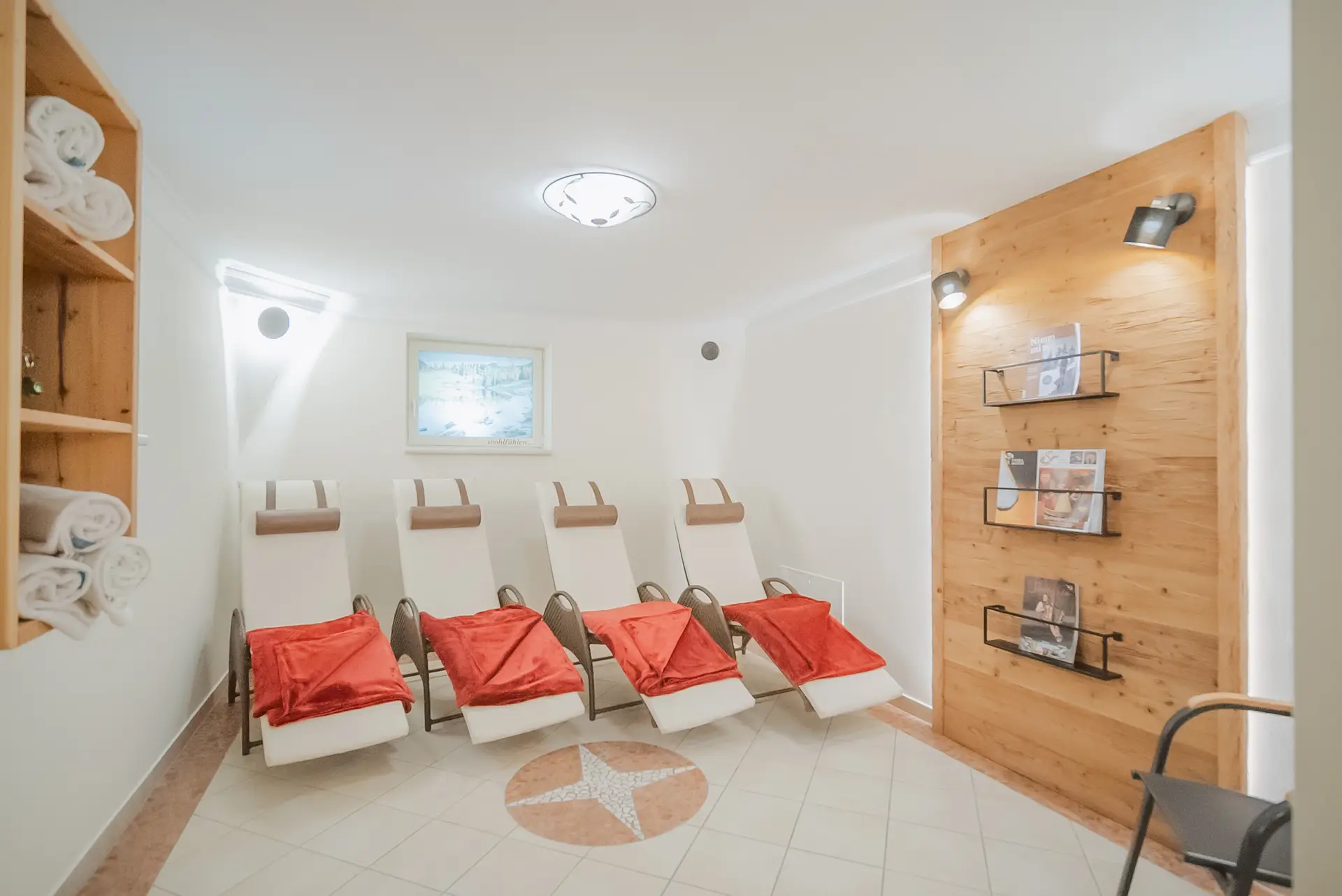 Ein Wellnessraum mit fünf Liegestühlen, jeder mit einer roten Decke, beige-weißen Wänden, einem Holzwandabschnitt mit Regalen für Zeitschriften, Handtuchhaltern auf der linken Seite und einer Deckenleuchte. Dieser gemütliche Ruheraum bietet eine ruhige Atmosphäre, die an einen erholsamen Urlaub am Bauernhof erinnert.